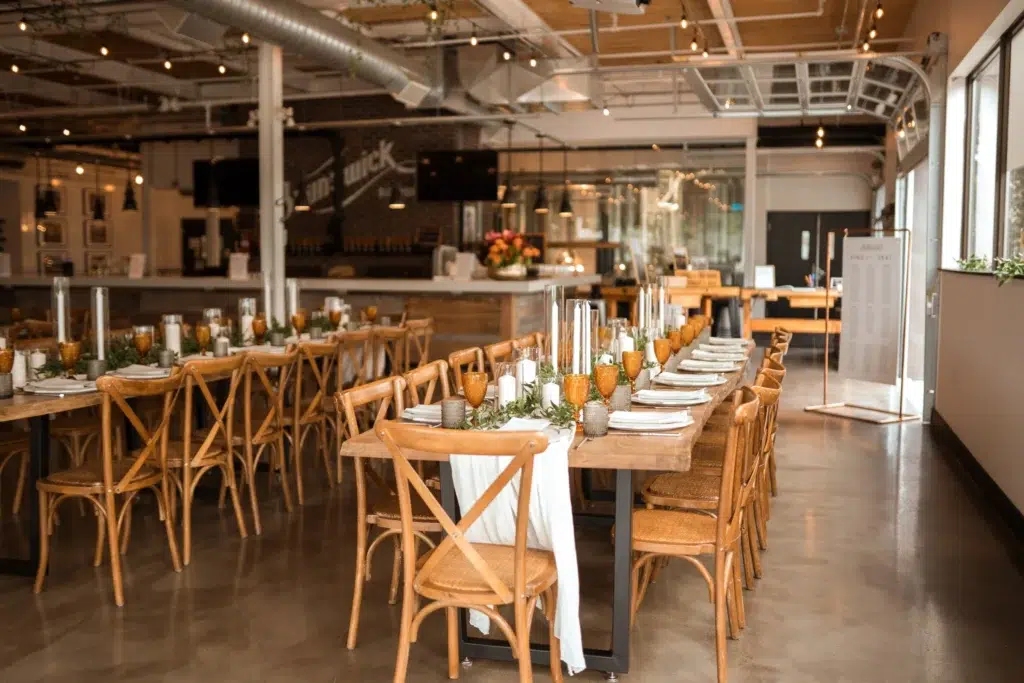 A modern restaurant with wooden chairs and long tables set with plates, glasses, candles, and greenery. Warm lighting and industrial décor create a welcoming atmosphere. The space is ready for guests.