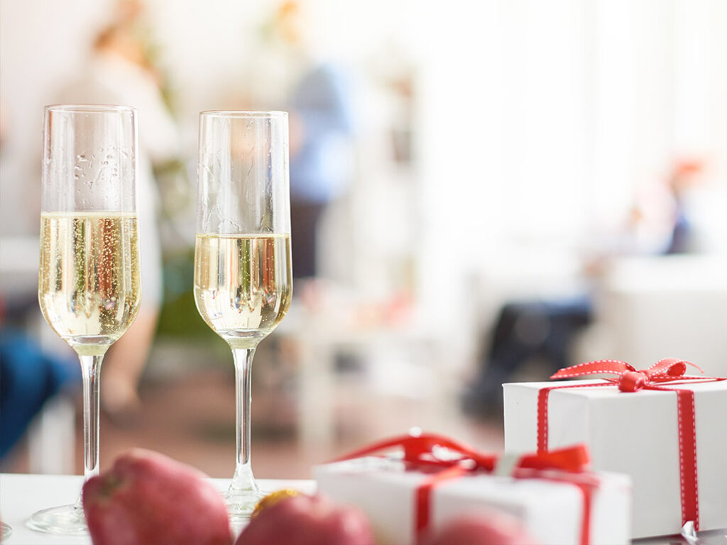 Two glasses of champagne on a table next to red apples and small white gift boxes with red ribbons, creating a festive atmosphere—perfect for a holiday catering event.