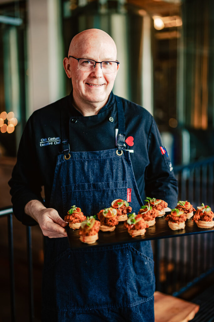 A smiling chef in a dark apron and chef's coat holds a tray of gourmet appetizers topped with greens, standing indoors with a blurred background—perfect inspiration for your next catering calendar event.