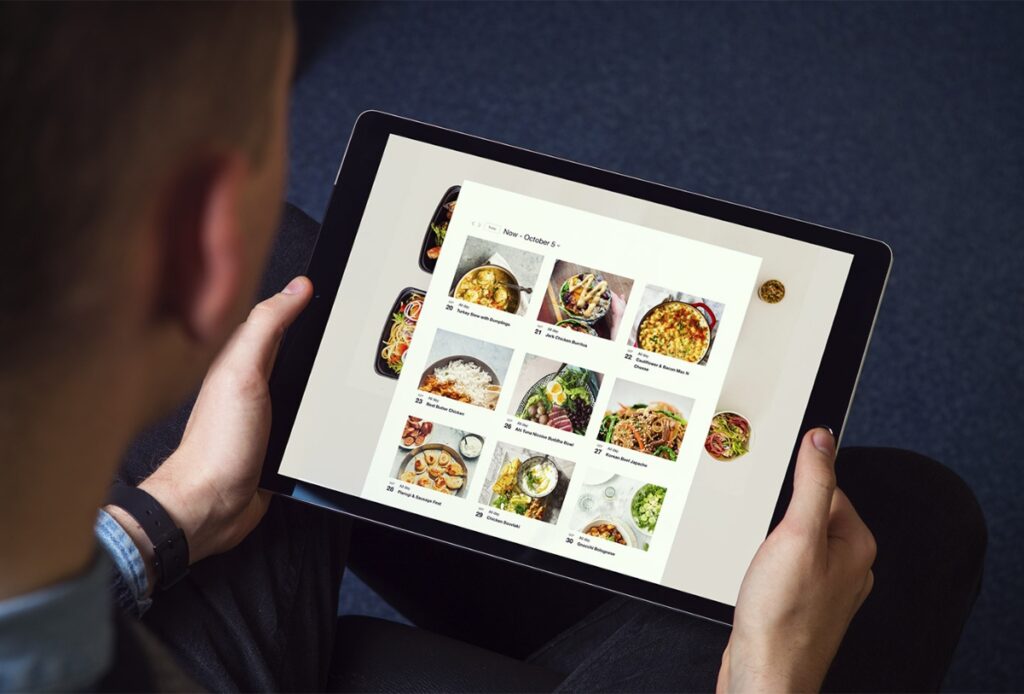 A person holds a tablet displaying a recipe website with a grid of food photos and dish names, viewed from above—perfect inspiration for office catering—set against a dark, textured background.