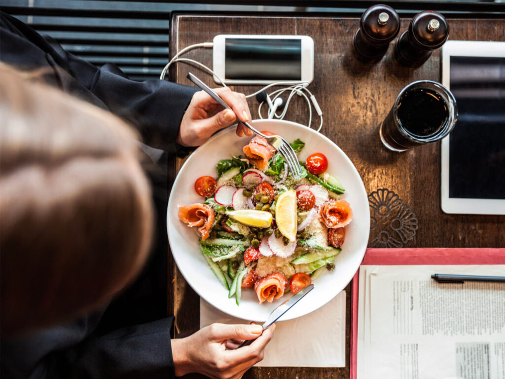 A person enjoying a fresh salad with smoked salmon, lemon, radish, cucumber, and cherry tomatoes at a table set for office catering with a drink, smartphone, earphones, tablet, and an open book or document.