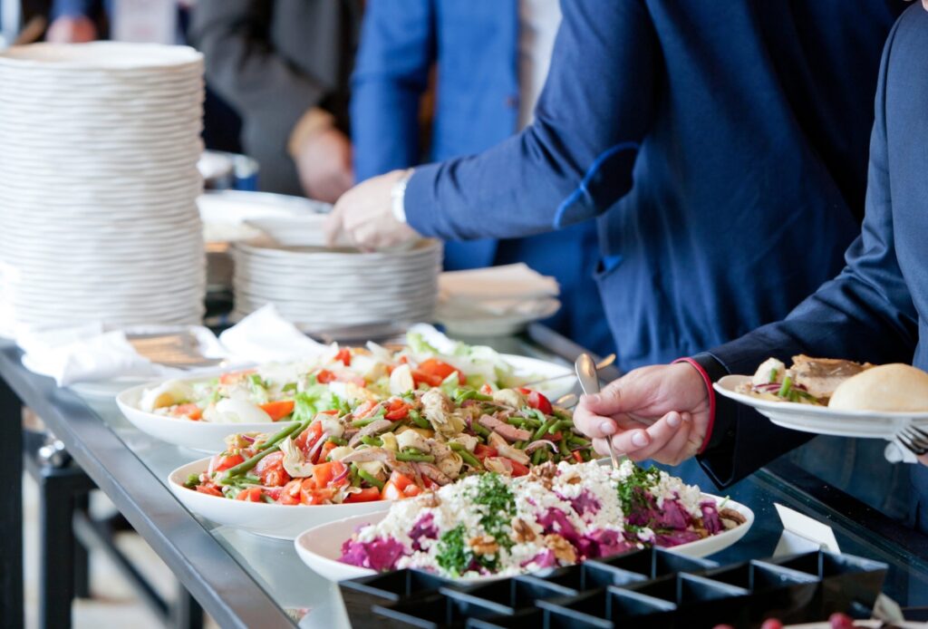 People in formal attire serving themselves food from an office catering buffet table with plates of salad, vegetables, and other dishes, and a stack of white plates nearby.