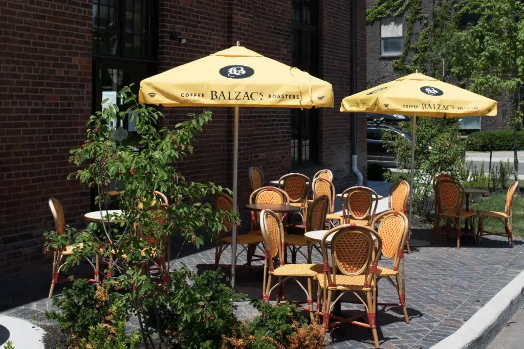 Outdoor café patio with yellow umbrellas labeled "Balzac's Coffee Roasters," featuring empty wicker chairs and tables on a stone pavement, surrounded by greenery and set against a brick building.