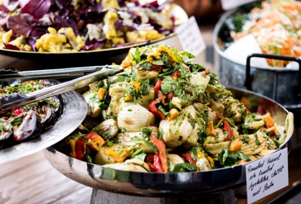 A large bowl of colorful roasted vegetables, including artichokes, peppers, and fresh herbs, sits on a buffet table alongside other assorted salads and dishes—perfect for vibrant office catering events.