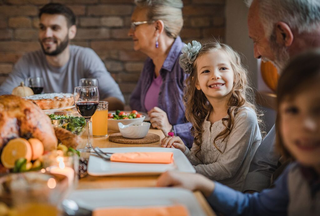 A smiling young girl sits at a dinner table with family members of various ages, surrounded by delicious food and drinks from thanksgiving catering, creating a warm and joyful atmosphere.