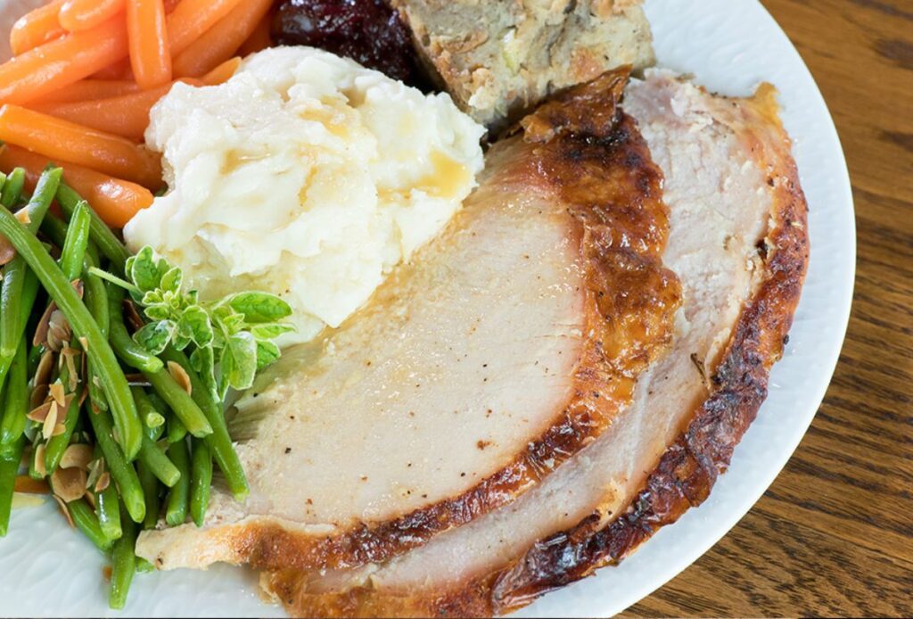 A plate with slices of roasted turkey, mashed potatoes with gravy, green beans with almonds, glazed carrots, stuffing, and cranberry sauce—perfect for memorable holiday catering.