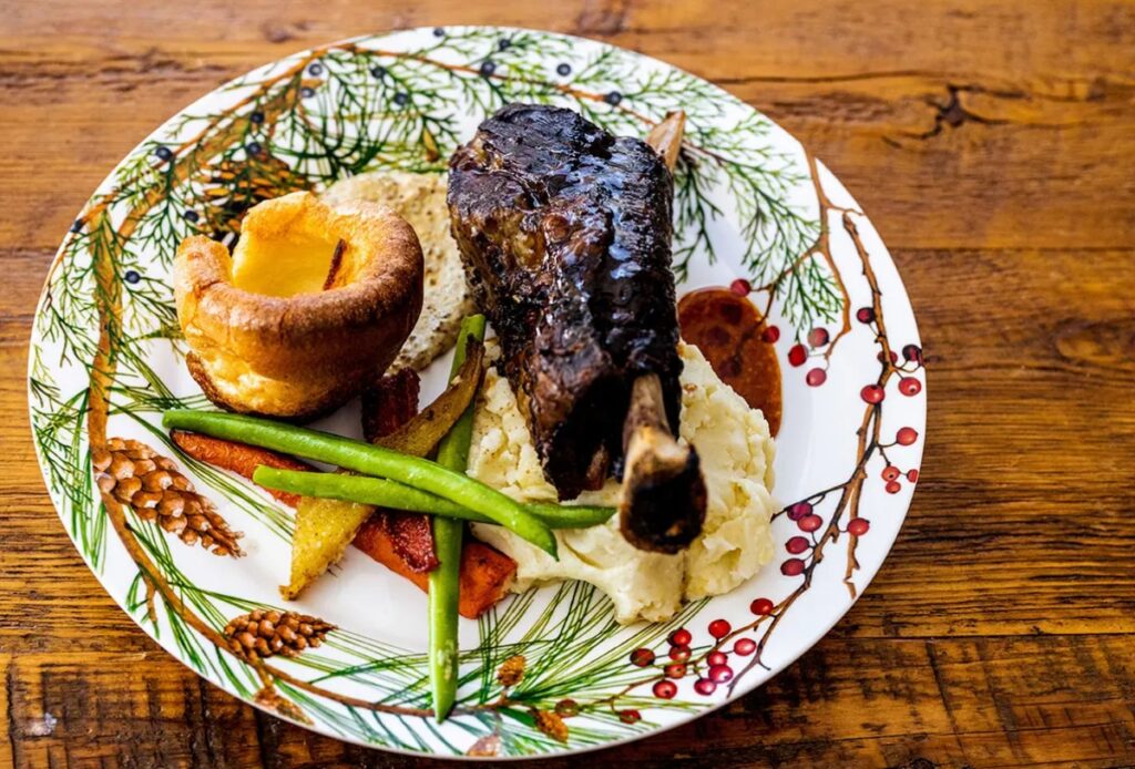 A festive plate for holiday party catering features roast meat on mashed potatoes, Yorkshire pudding, sliced carrots, green beans, and bread, all served on a pine cone and berry decorated plate atop a wooden table.