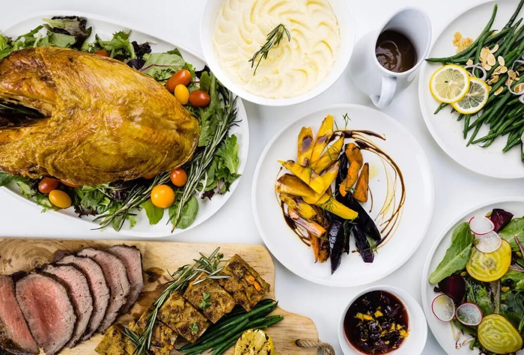 A festive table set for holiday party catering features roasted turkey, mashed potatoes with gravy, roasted vegetables, green beans with almonds, a radish and lemon salad, sliced roast beef, stuffing, and cranberry sauce.