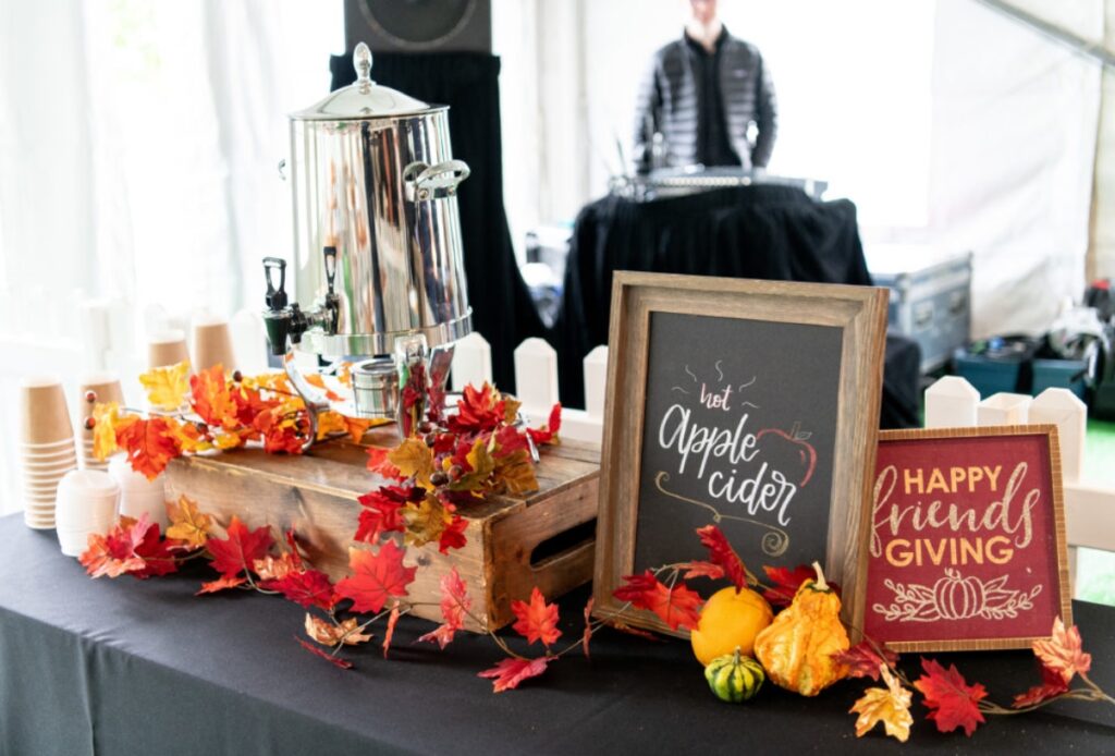 A festive table for holiday party catering features a metal drink dispenser, paper cups, autumn leaves, small gourds, and framed signs reading "Hot Apple Cider" and "Happy Friendsgiving." A person stands in the blurred background.