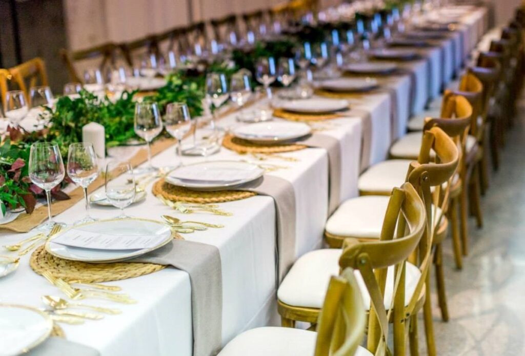 A long banquet table set for a formal event with white tablecloths, elegant plates, wine glasses, gold cutlery, woven placemats, and green leafy centerpieces offers the perfect setting for memorable holiday party catering, surrounded by wooden chairs.