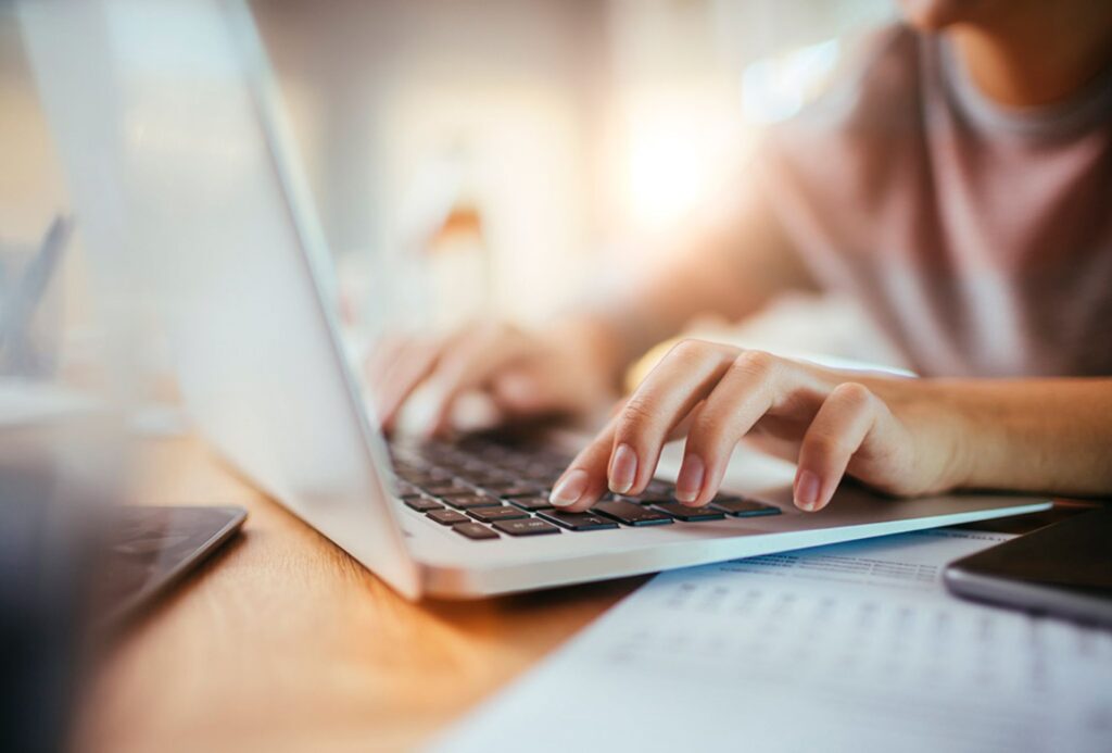Close-up of a person’s hands typing on a laptop keyboard, with papers and a pen nearby—perhaps planning thanksgiving catering—in a bright indoor setting.