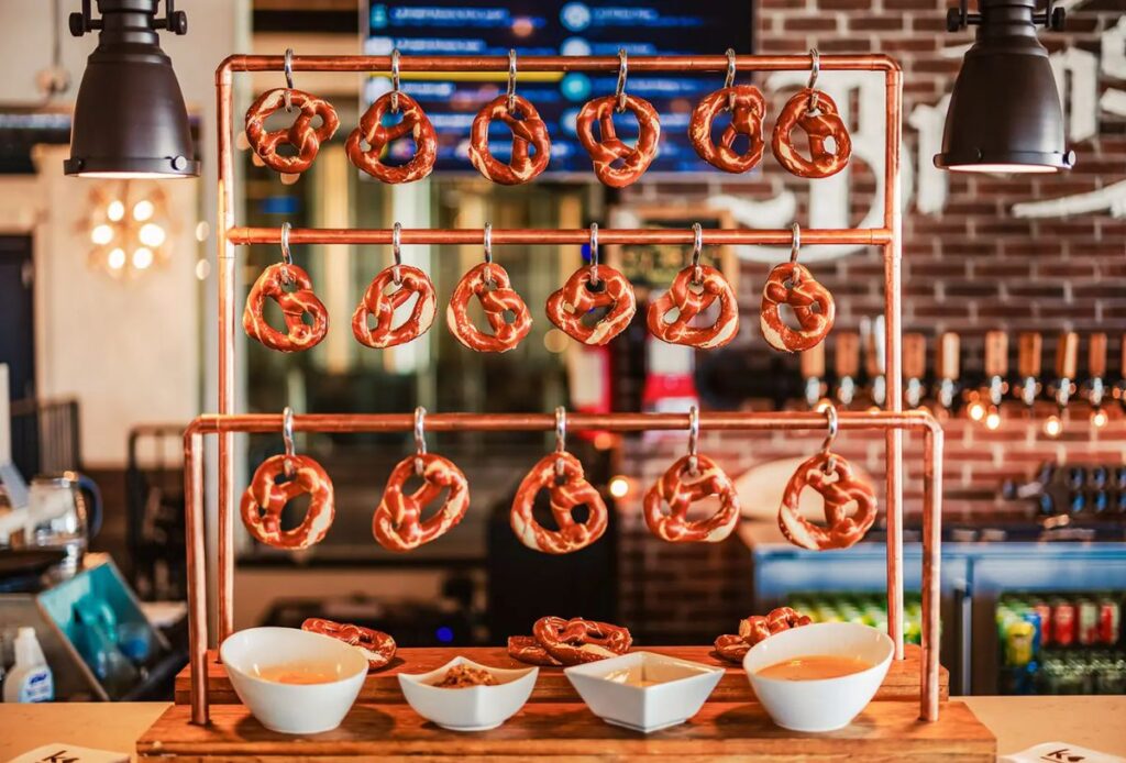 Soft pretzels hang on metal hooks from a copper rack, with three white bowls of dips and snacks below—an inviting addition to any holiday catering spread. In the background, a bar area features bottles and a rustic brick wall.