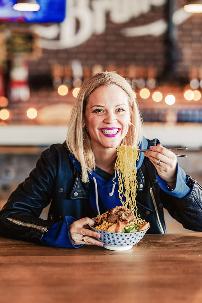 A smiling woman with blonde hair holds up a serving of noodles with chopsticks over a bowl of ramen while sitting at a wooden table, enjoying a meal prepared by a renowned Toronto catering company.