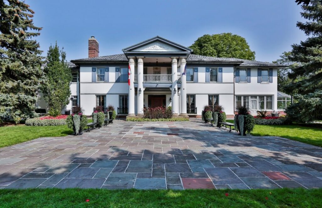 A large white two-story mansion with columns at the entrance, a balcony, and a wide stone driveway, surrounded by green lawns, trimmed bushes, trees, and colorful flower beds.