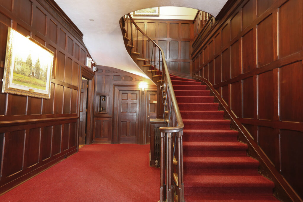 A grand staircase with red carpet and curved wooden banister leads to an upper floor. The walls are paneled with dark wood and decorated with framed paintings. Warm lighting brightens the elegant hallway.