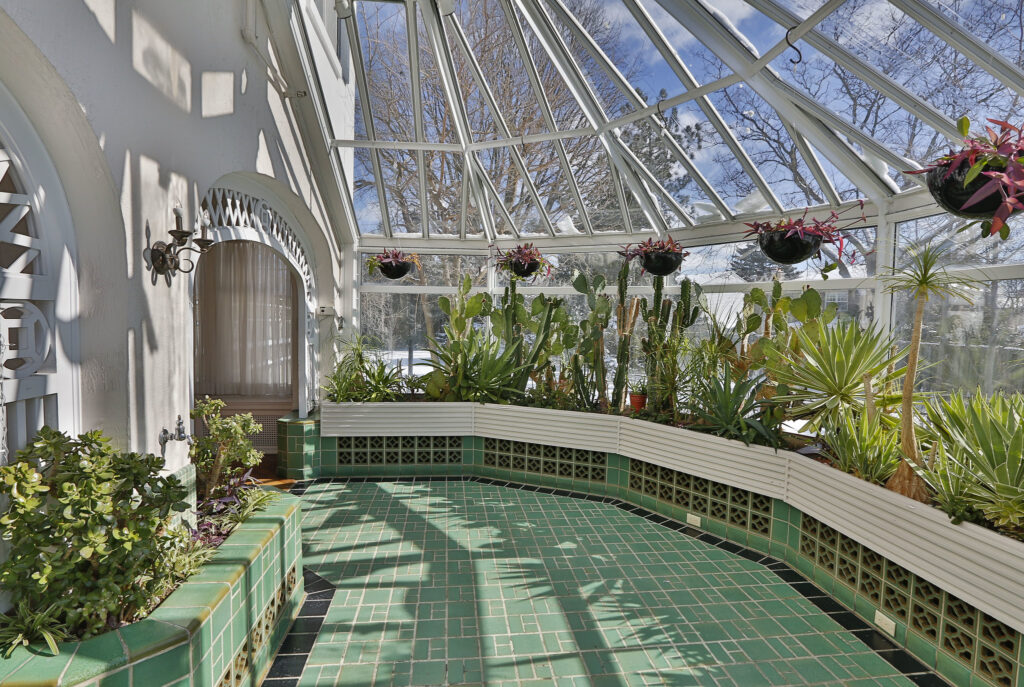 A bright sunroom with a glass ceiling and walls, featuring green-tiled floors and built-in planters filled with various cacti and potted plants. Hanging planters decorate the space, with trees visible outside.