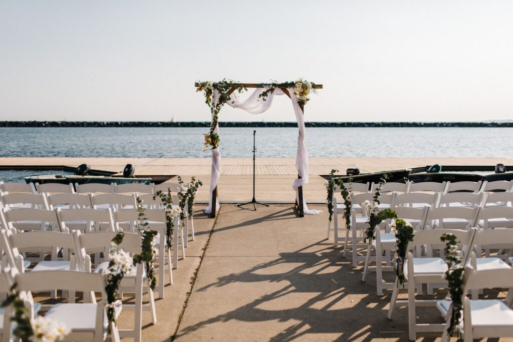Rows of white chairs face a floral-decorated arch and microphone stand on a dock overlooking calm water, set up for an outdoor wedding ceremony on a sunny day.