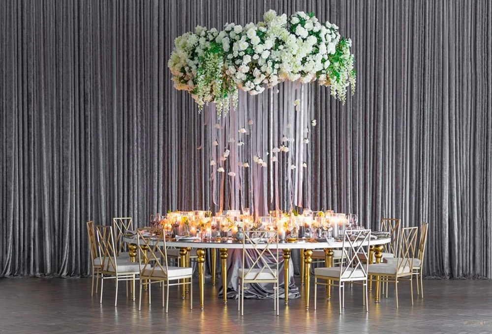 A round table with gold chairs is elegantly set with candles and glassware. Above the table, white and green flower arrangements hang from the ceiling against a backdrop of silver-gray curtains.