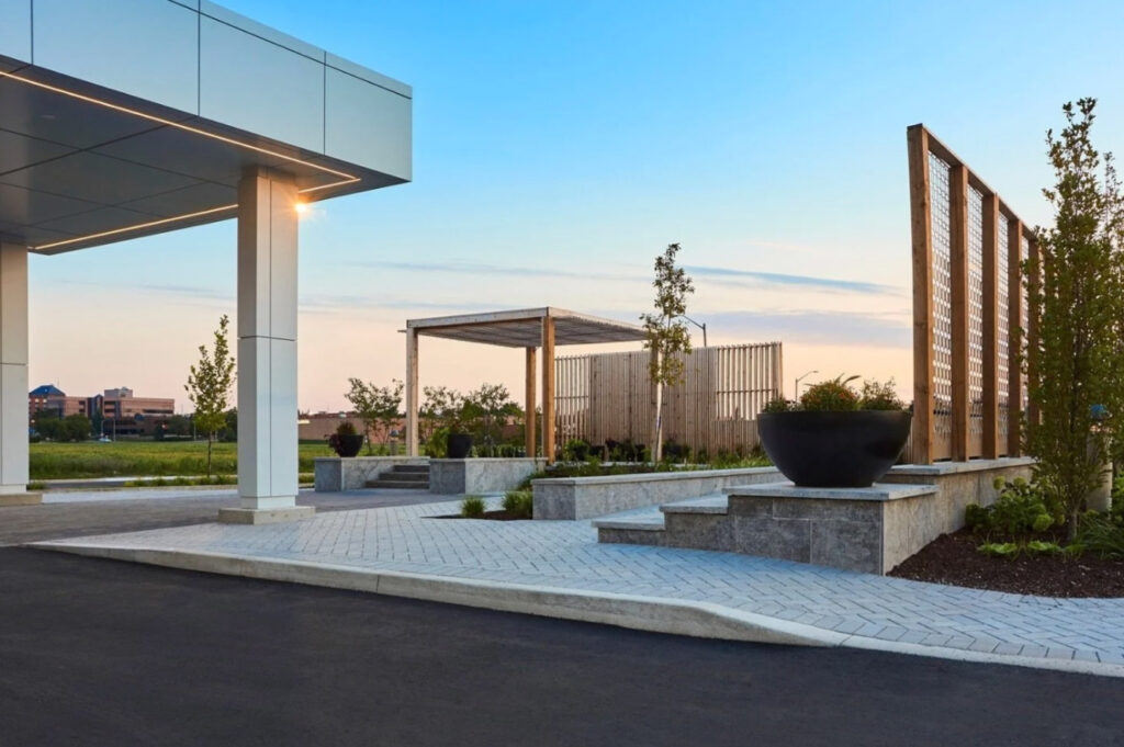 Modern outdoor area with geometric landscaping, large planters, young trees, and wooden trellises. The space features stone walkways, raised beds, and a building entrance canopy, set against a clear sky at dusk.