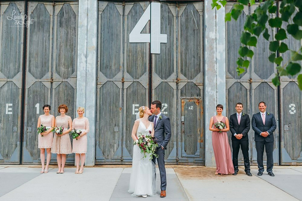 A bride and groom kiss in front of a large industrial door marked with the number 4, flanked by three bridesmaids in light pink dresses and three groomsmen in dark suits. Green leaves frame the top right corner.