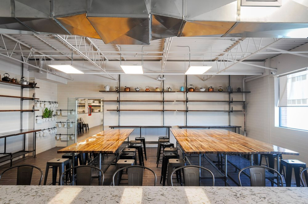 A spacious, modern kitchen with polished wooden tables, black metal chairs, open shelves with kettles and plants, white tiled walls, large windows, and exposed ductwork on the ceiling.
