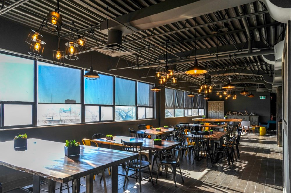 A modern, sunlit dining area with wooden tables and black chairs, industrial-style lighting, large windows with blue shades, potted plants, and exposed ceilings.