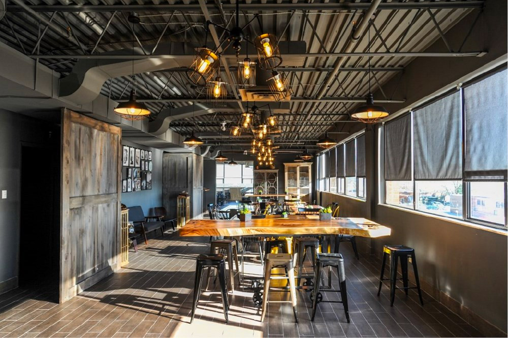 A modern, industrial-style room features large windows with rolled-up blinds, a wooden table surrounded by metal stools, exposed ceiling pipes, and hanging Edison bulb lights. The space is filled with natural sunlight.