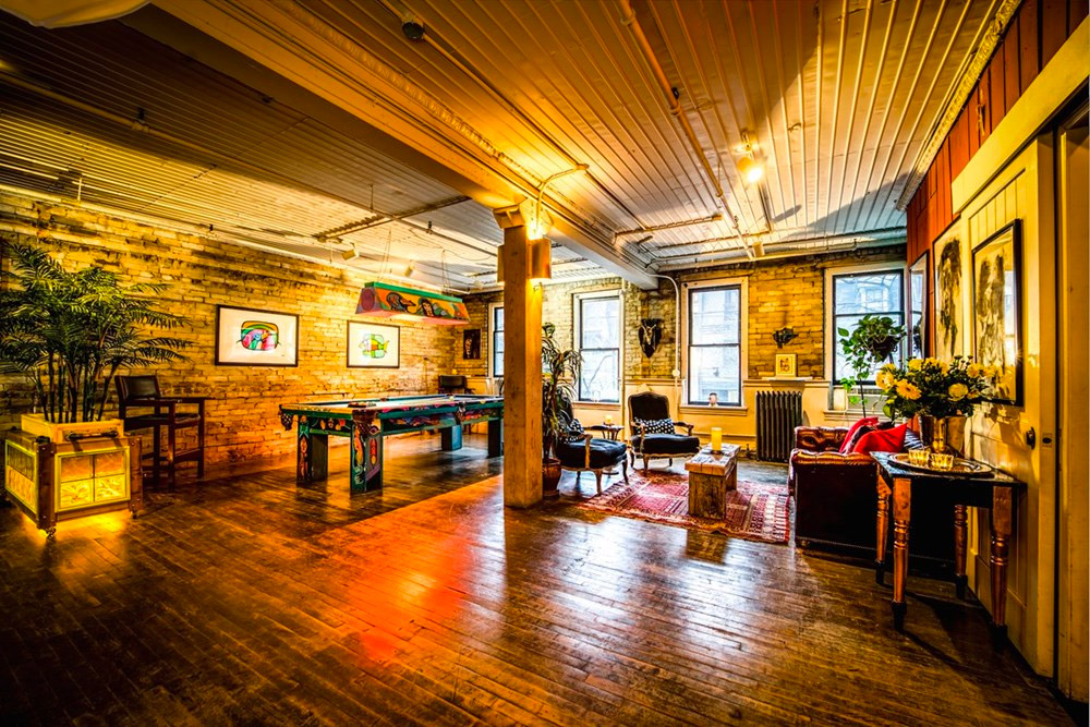 A spacious, warmly lit loft with exposed brick walls, wooden floors, eclectic furniture, plants, colorful artwork, a pool table, and large windows letting in natural light.