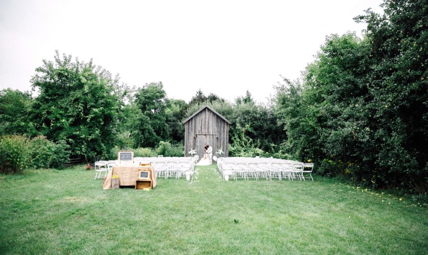 An outdoor wedding setup with rows of white chairs facing a small rustic wooden barn, surrounded by green trees and grass. A bride in white stands at the barn entrance. A wooden table stands to the left.