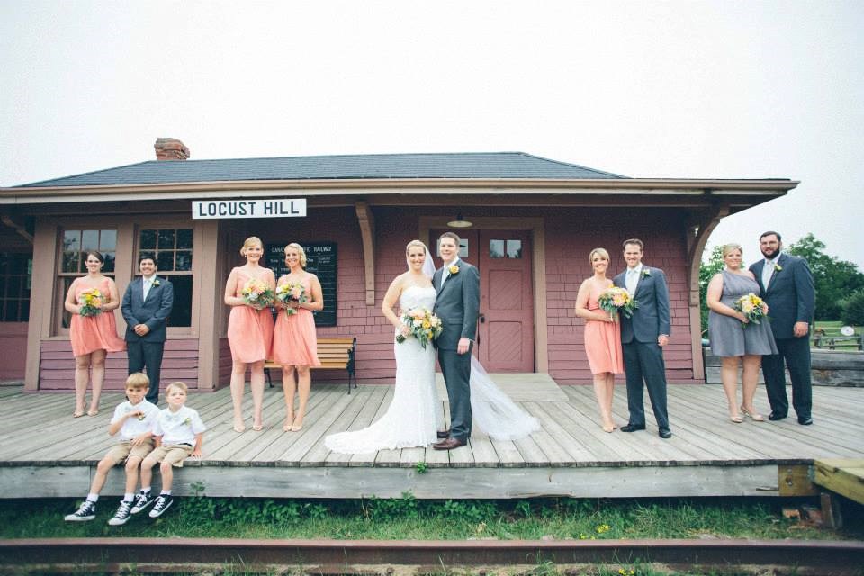 A wedding party poses on a wooden platform in front of a building labeled “Locust Hill.” The bride and groom stand in the center, surrounded by bridesmaids in peach dresses and groomsmen in gray suits. Two children sit on the steps.