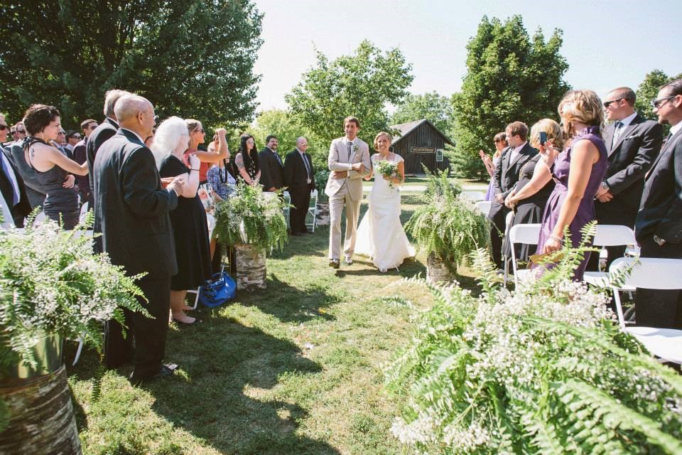 A bride in a white dress walks down an outdoor aisle on green grass, escorted by a man in a tan suit. Guests stand and watch, and large ferns decorate the aisle. Trees and a wooden building are visible in the background.