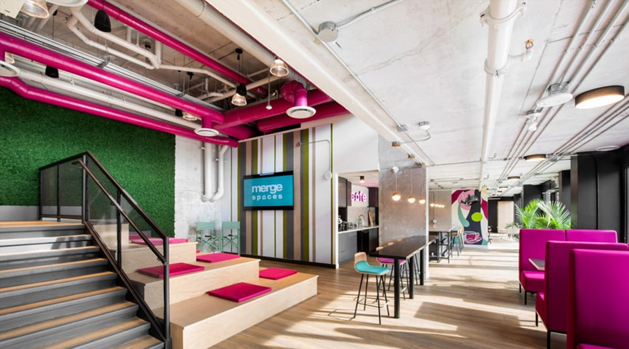 A modern, open-concept coworking space with bright pink accents, tiered seating with cushions, a staircase, communal tables, greenery on the wall, and exposed ceilings with visible pipes.