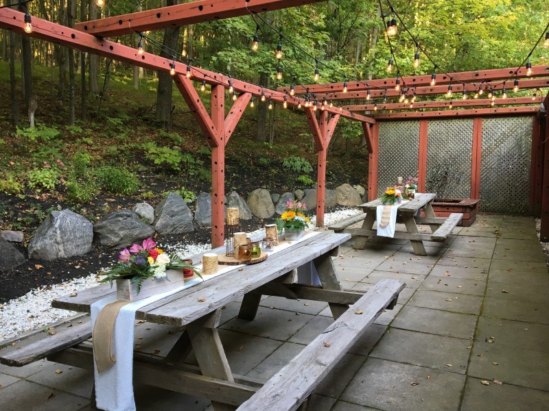 Outdoor patio with two wooden picnic tables, decorated with flowers and candles, under a wooden pergola with string lights. Surrounded by trees and rocks, creating a cozy, rustic setting.