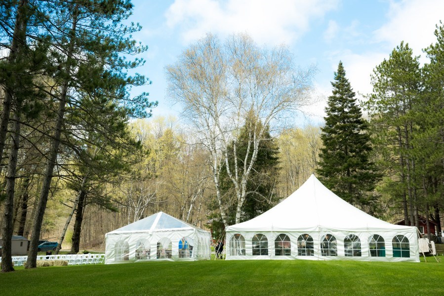 Two white event tents are set up on a grassy lawn surrounded by tall trees, with a partly cloudy sky overhead. One tent is smaller and square, the other is larger and round with arched windows.