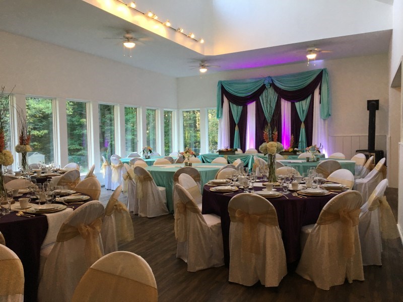 A decorated banquet hall with round tables covered in white cloths, gold and purple accents, and elegant place settings. A stage with teal and purple drapes is at the front, and large windows offer views of greenery outside.