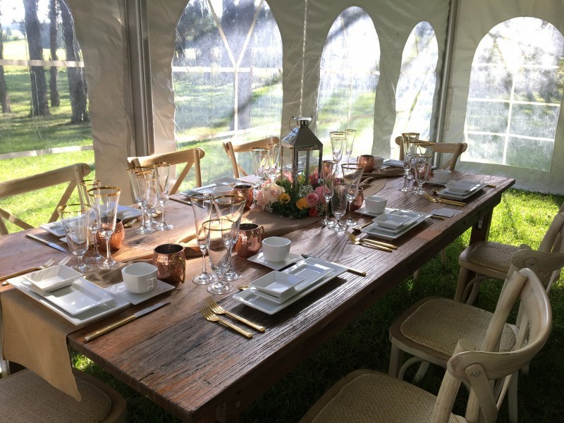 A rustic wooden table set for a formal outdoor meal with white plates, gold utensils, glassware, copper mugs, and a flower centerpiece under a tent with natural light.