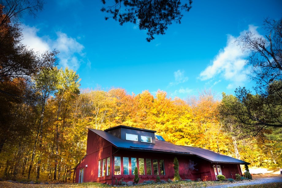 A red house with large windows sits in front of vibrant autumn trees with golden-yellow leaves under a bright blue sky with scattered clouds.