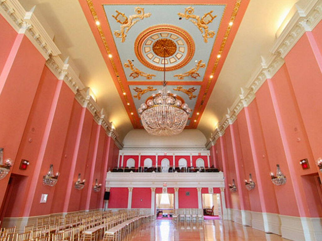A grand ballroom with pink walls, gold and white trim, an ornate blue and gold ceiling, and a large crystal chandelier. Rows of empty chairs line the polished wood floor, and balconies overlook the room.
