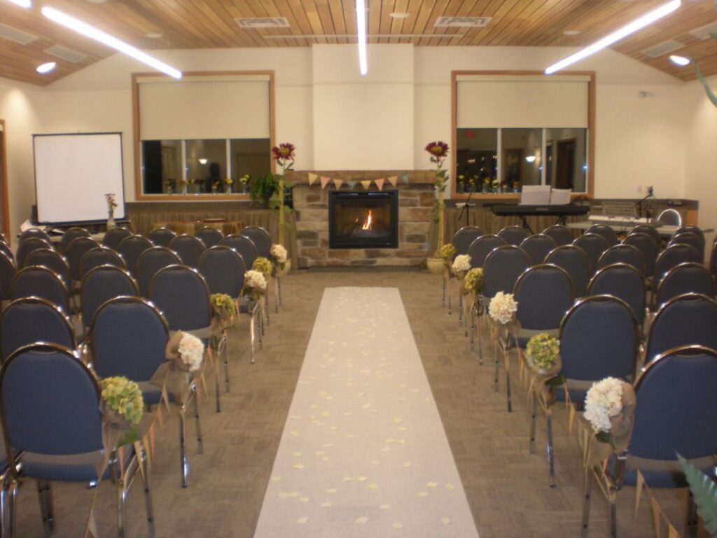 A room set up for a wedding ceremony features rows of chairs with white floral decorations along the aisle, a white runner leading to a fireplace, and soft lighting from the ceiling.
