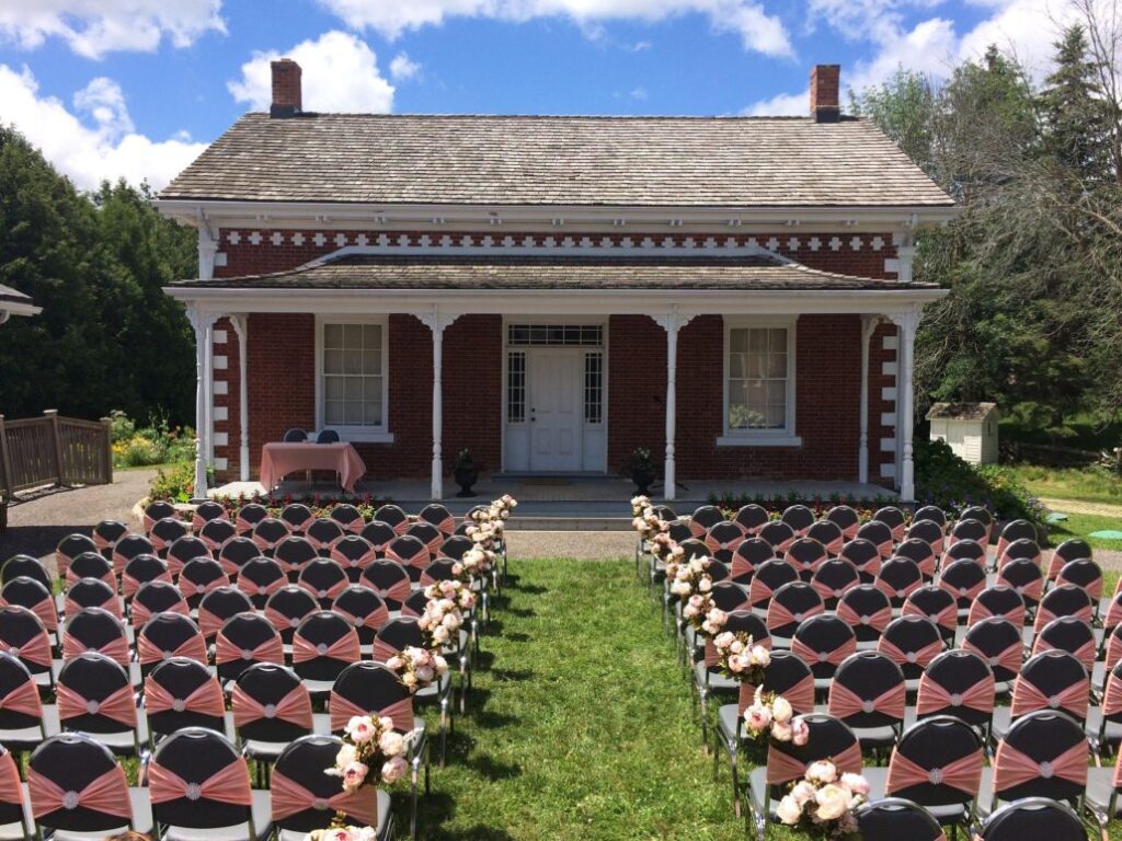 Rows of decorated chairs are arranged on a lawn facing a red brick house with white trim, set up for an outdoor wedding ceremony under a partly cloudy sky.