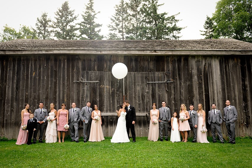 A wedding party stands in front of a rustic wooden barn. The bride and groom kiss under a white balloon, surrounded by bridesmaids in pink dresses and groomsmen in grey suits, all smiling on green grass.