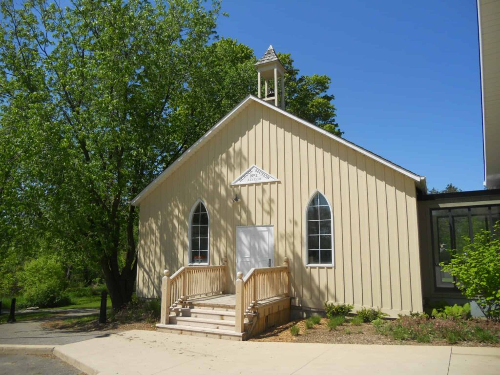 A small, cream-colored chapel with arched windows, a steeple, and a wooden ramp, surrounded by trees and greenery under a clear blue sky.