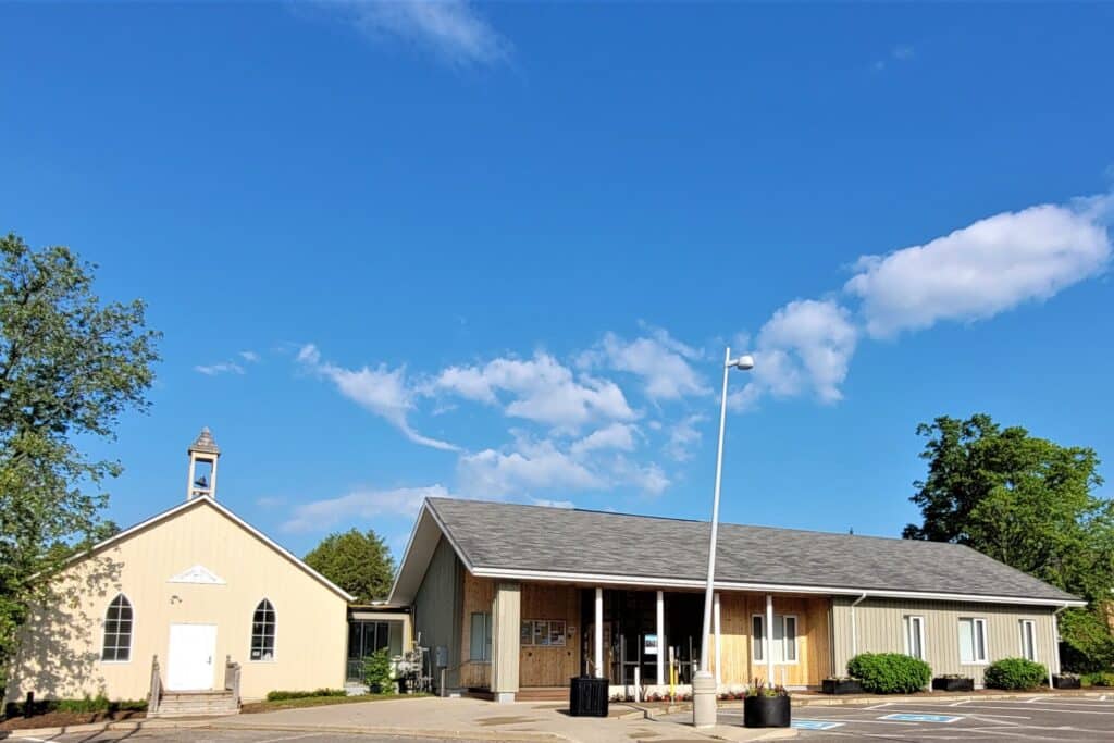 A small beige church with a steeple stands next to a larger building with a covered entrance, both under a bright blue sky with scattered clouds. Trees and an empty parking lot surround the buildings.