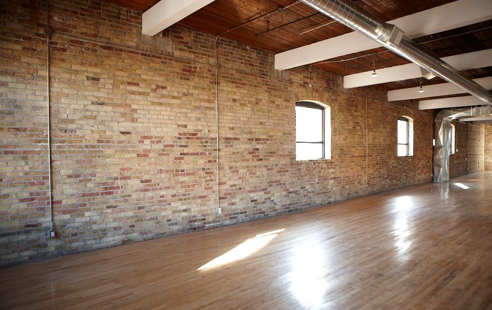 Sunlight streams into a spacious room with exposed brick walls, wooden floors, large windows, and industrial ceiling pipes, creating a warm, open, and loft-like atmosphere.