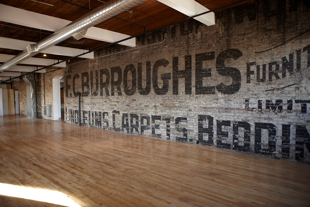 A spacious loft with exposed brick walls, visible ductwork on the ceiling, and wooden floors. The brick wall features large, faded black text advertising "F.C. BURROUGHES" and "Carpet & Bedding.