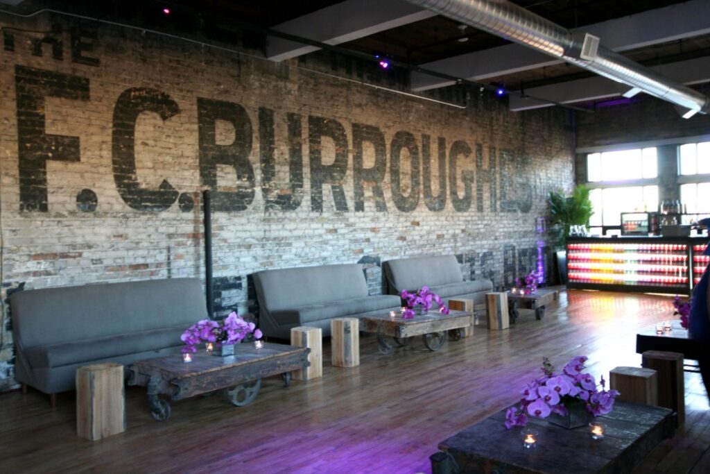A stylish lounge with gray couches, rustic wooden tables topped with purple flowers, and exposed brick walls featuring large faded text. Colorful lights illuminate the bar at the back, creating a modern industrial ambiance.