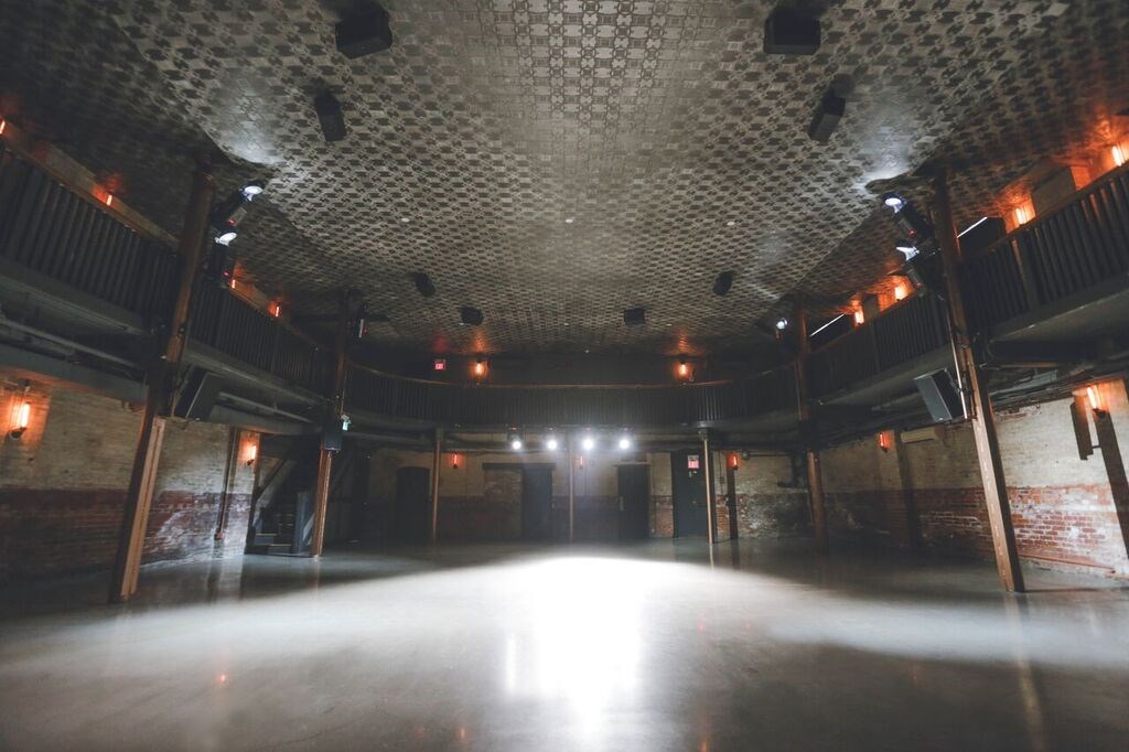 An empty, spacious event hall with a patterned tin ceiling, exposed brick walls, soft wall lighting, and a balcony level running along the sides. Bright lights illuminate the polished floor from the back of the room.