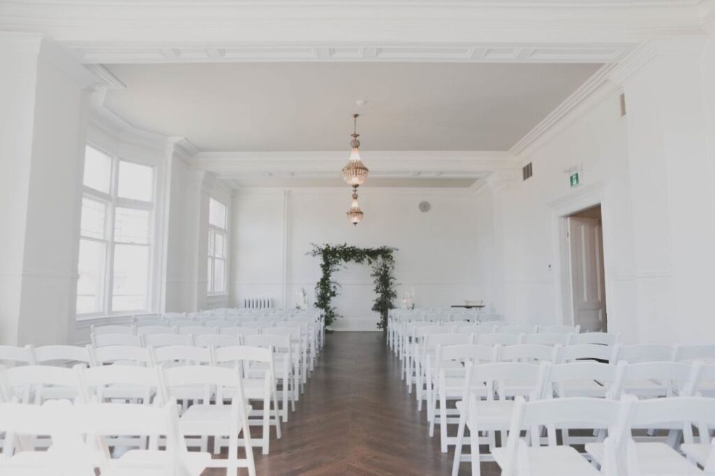 A bright, airy room set up for a wedding ceremony with rows of white chairs facing a simple greenery arch at the front. Chandeliers hang from the ceiling, and large windows let in natural light.
