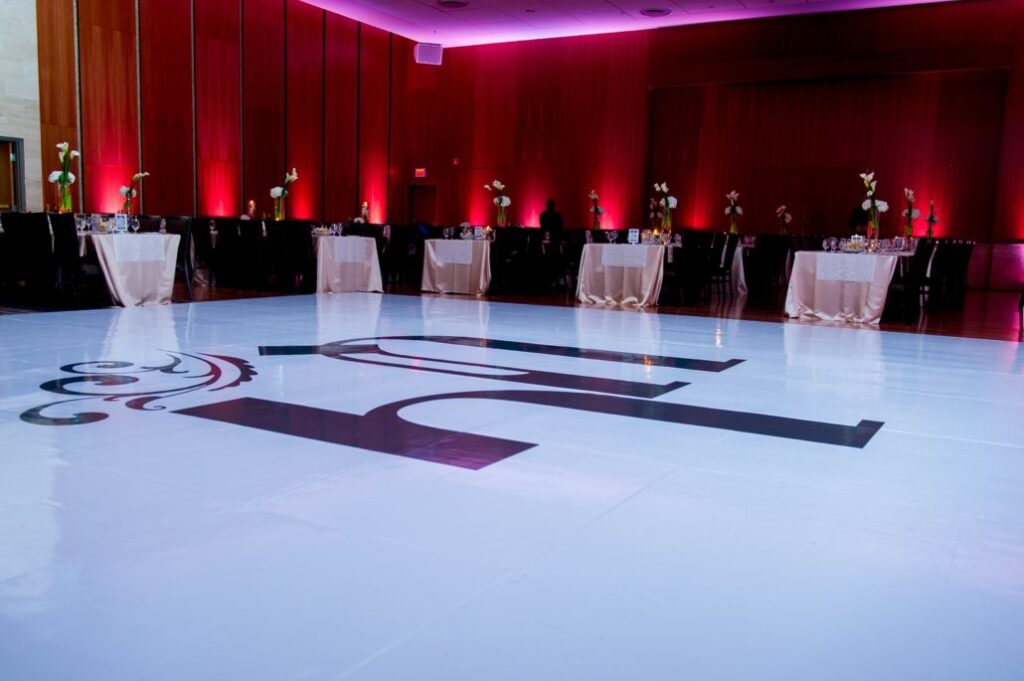 A spacious, elegant event hall with a glossy white dance floor featuring a large stylized letter "H," surrounded by tables draped in white linens and decorated with flowers, and purple ambient lighting along the walls.