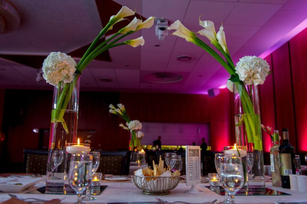 Elegant banquet table with tall glass vases holding white flowers and calla lilies, surrounded by candles, wine bottles, glasses, and snacks; the room is lit with pink and purple ambient lighting.